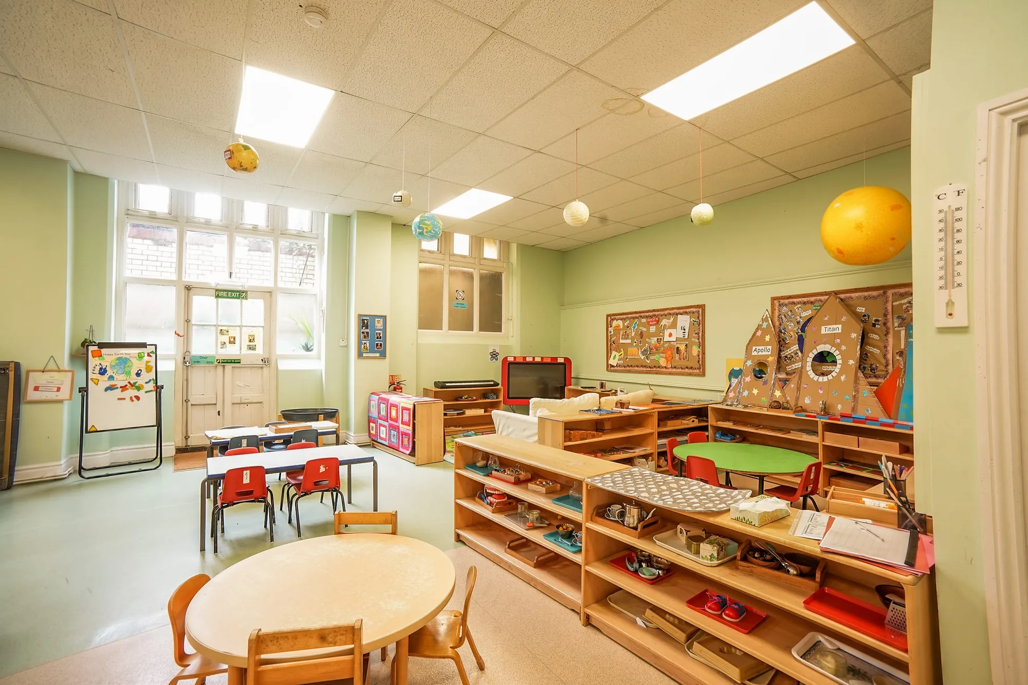 A wide view of the Angel Montessori classroom with low shelves, round tables and a fire-exit door