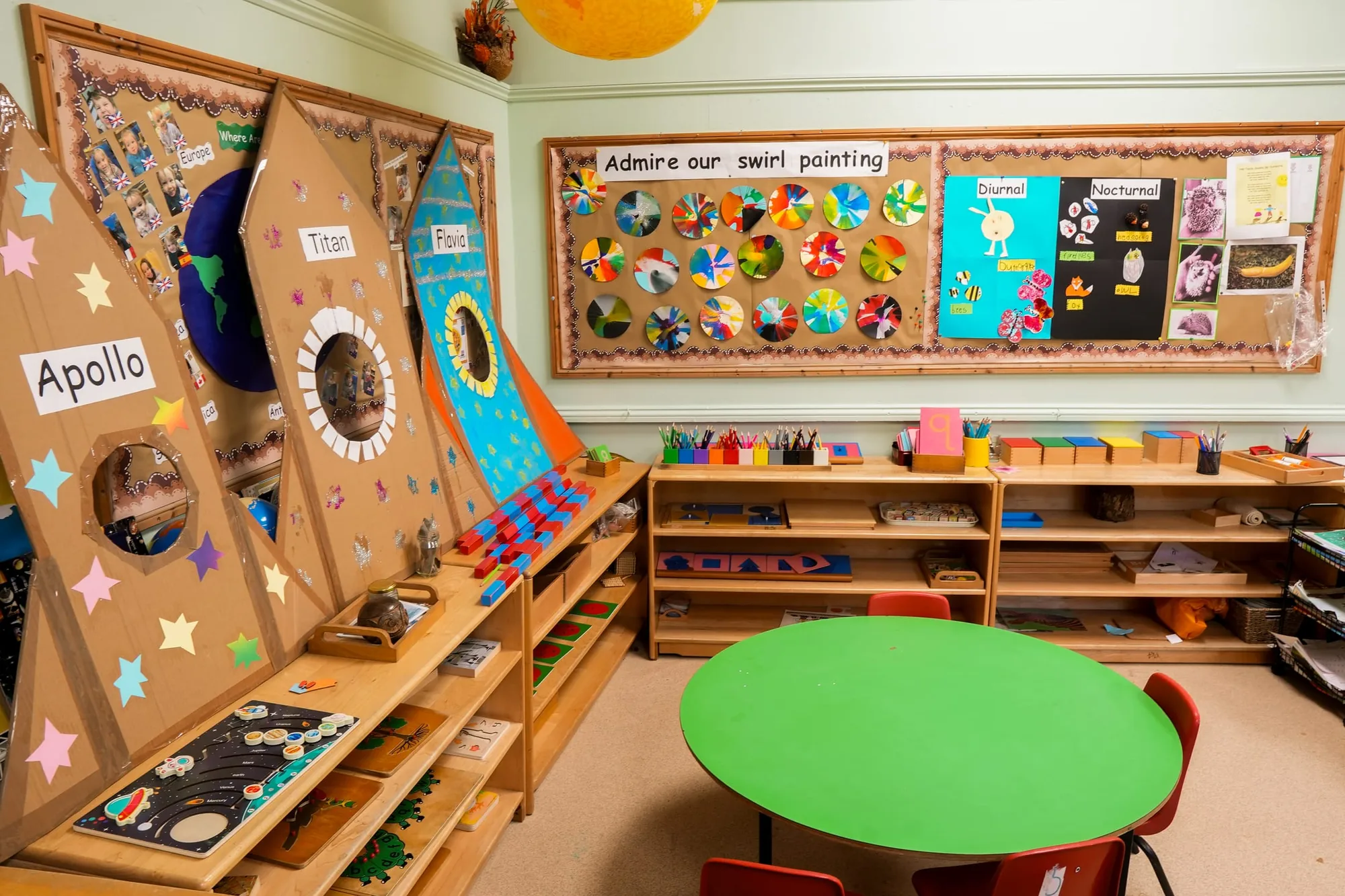 Cardboard rocket displays above shelves of Montessori materials and a green table