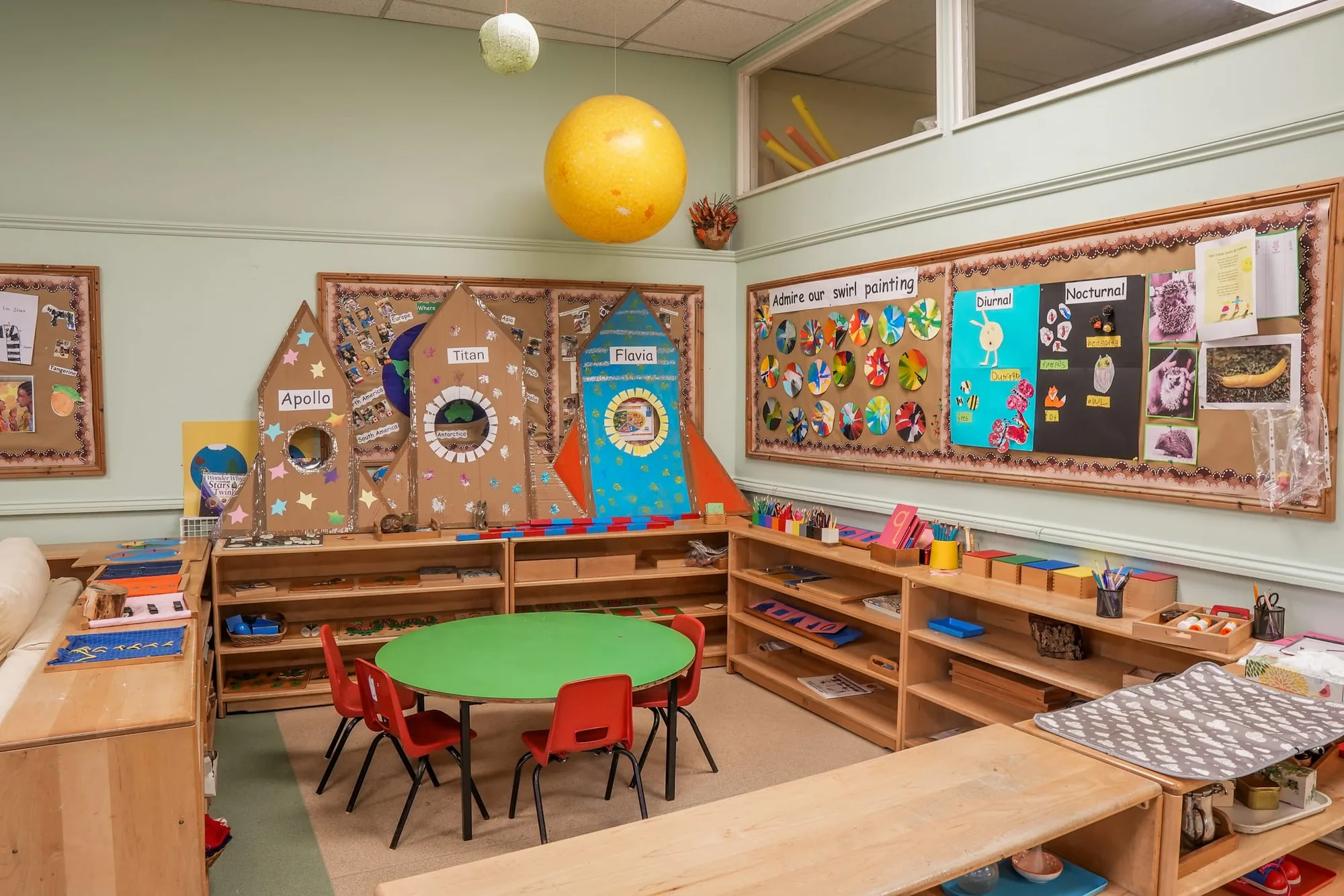 Montessori materials on low shelves around a green table, with children’s swirl paintings